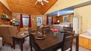 A cozy cabin interior with a wooden dining table, chairs, a sofa, and a separate sleeping area with a bed.