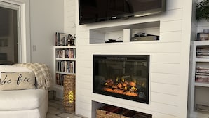 A living room with a fireplace, a bookshelf, and a TV mounted on the wall. - Beachfront Baby💛 Beautiful & New!🌟The only way to stay @ the beach❣️Spacious  (Dewey Beach)