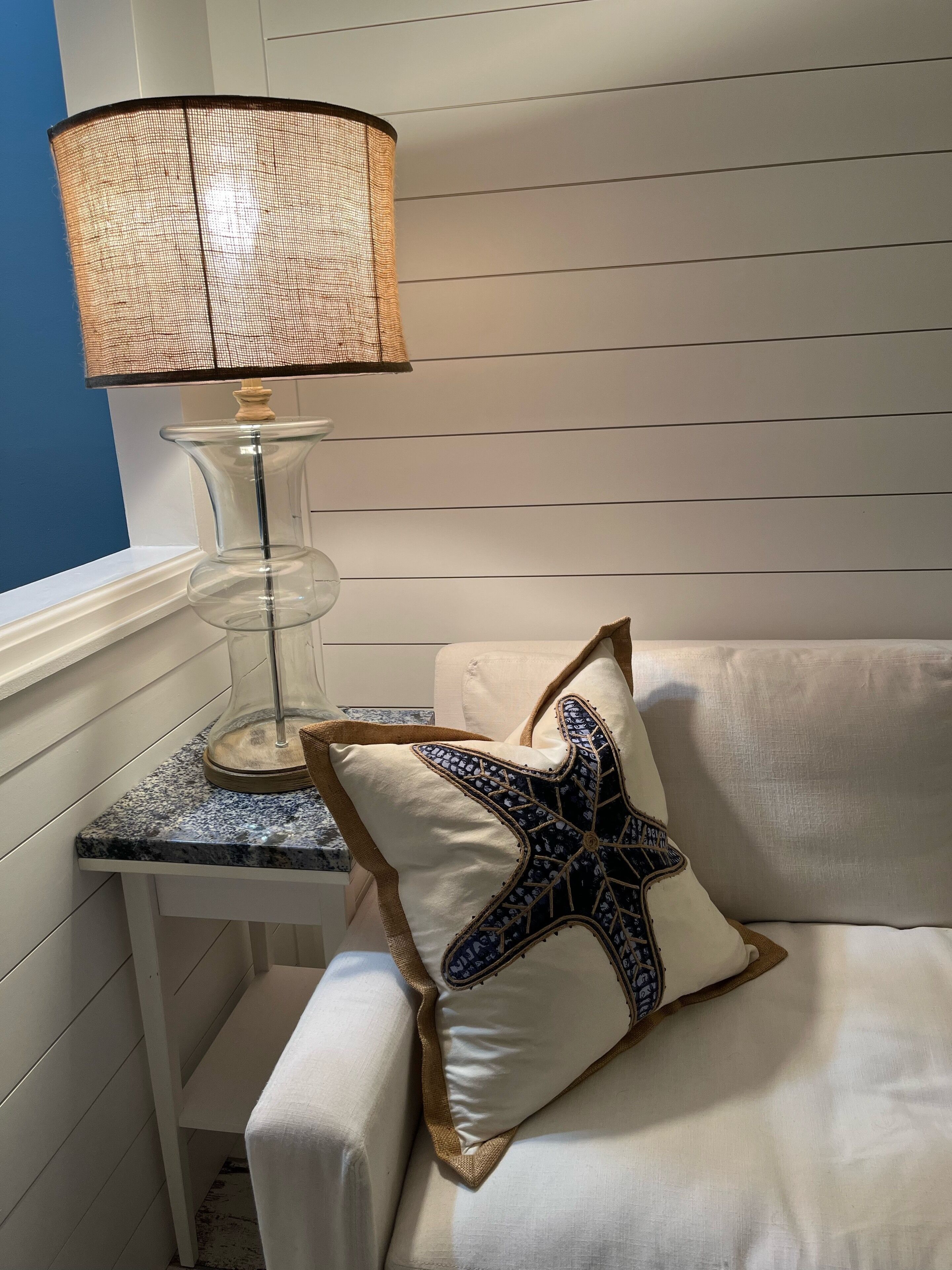 Un coin de pièce avec un banc blanc rembourré, une lampe en verre transparent dotée d’un abat-jour en osier, et un coussin orné d’un motif en forme d’étoile de mer.