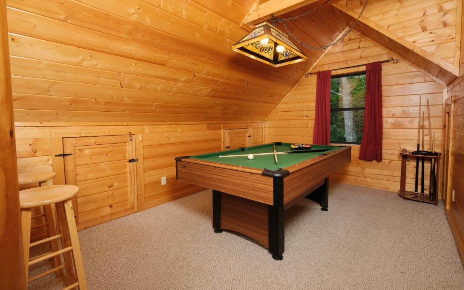 A wooden-paneled room with a pool table, a ladder, and a window with red curtains.