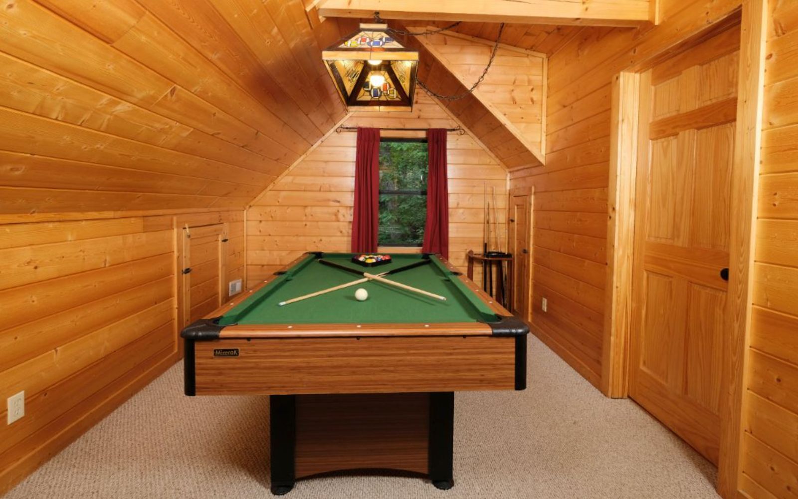 A pool table in a wooden room with a sloped ceiling and a hanging light fixture.