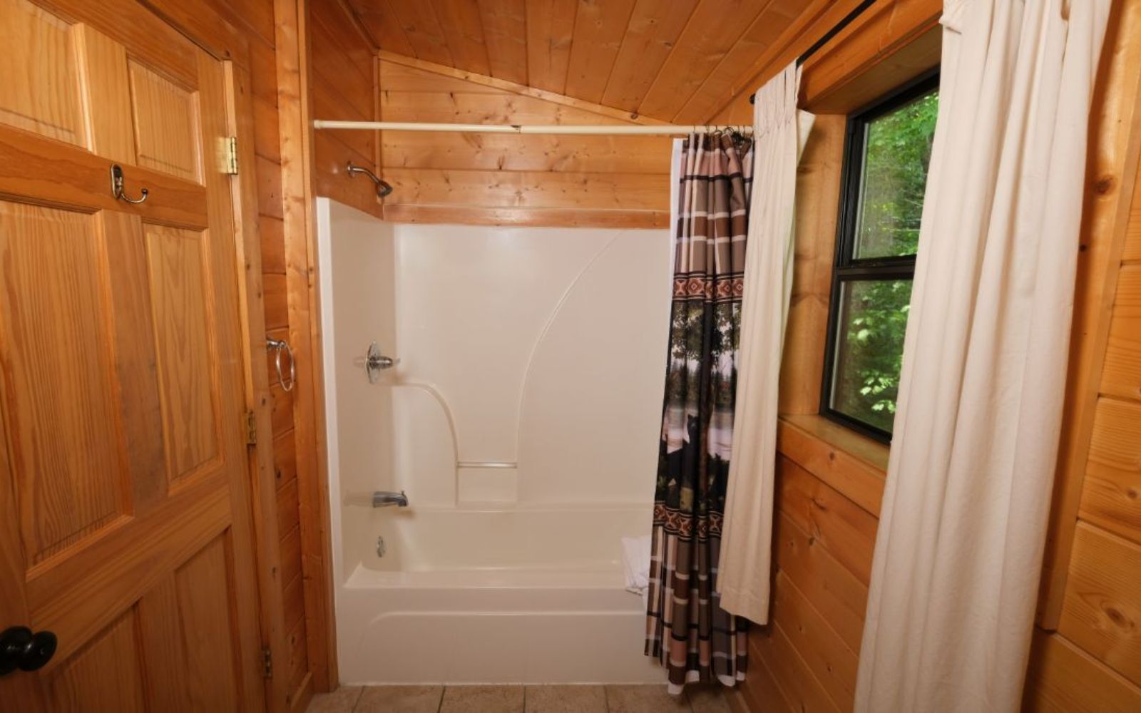 Baño de cabaña de madera con bañera, cortina de ducha y ventana con vistas a la vegetación.