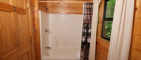 A wooden cabin bathroom with a bathtub, a shower curtain, and a window with a view of greenery.