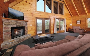 Cozy cabin with a stone fireplace, comfortable sofa, and flat-screen TV, offering views of the surrounding landscape from the balcony and large windows.