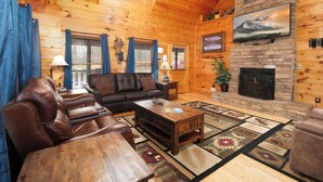 A cozy log cabin interior with a fireplace, leather sofa, and a large wooden table. - The Looney Bin - Hot Tub, Game Loft, Jetted Tub Suite & Wooded Views (Sevierville)