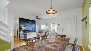 A spacious living area with a large wooden dining table, a sofa set, and a television mounted on the wall. - Moonsong is in a great location, is golf cart accessible and just a short drive (Port Aransas)