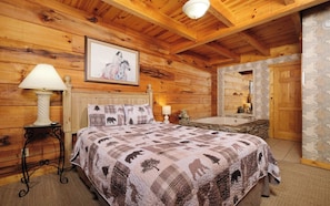 A cozy log cabin bedroom with a large bed, a nightstand with a lamp, a framed picture on the wall, and a stone fireplace.