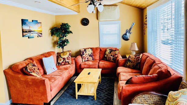 A cozy living room with a wooden ceiling, a ceiling fan, a large window with blinds, a wall clock, a guitar decoration, and a potted plant. - Upscale and Cozy Ocean Block Home w/ Free Parking, BBQ, Patio, Outdoor Shower (Seaside heights)