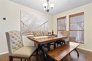 A dining room with a wooden table, four upholstered chairs, a chandelier, and large windows with blinds.