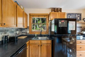 A kitchen with wooden cabinets, a black refrigerator, a window with a view of trees, and a coffee bar sign.
