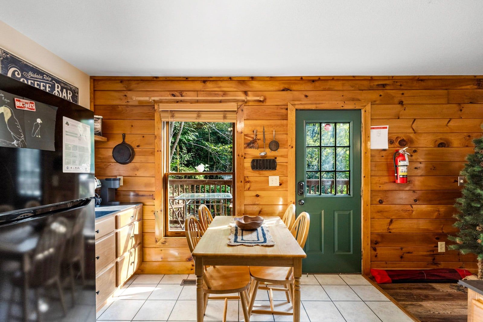 L’intérieur d’un chalet en bois comprend une table à manger, des chaises, un réfrigérateur, une porte verte et une fenêtre donnant sur un paysage verdoyant.