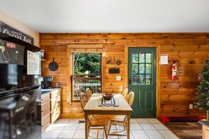 A wooden cabin interior with a dining table, chairs, a refrigerator, a green door, and a window overlooking greenery.