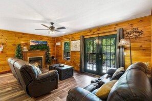 A cozy log cabin interior with a fireplace, leather seating, and a view through the glass doors.