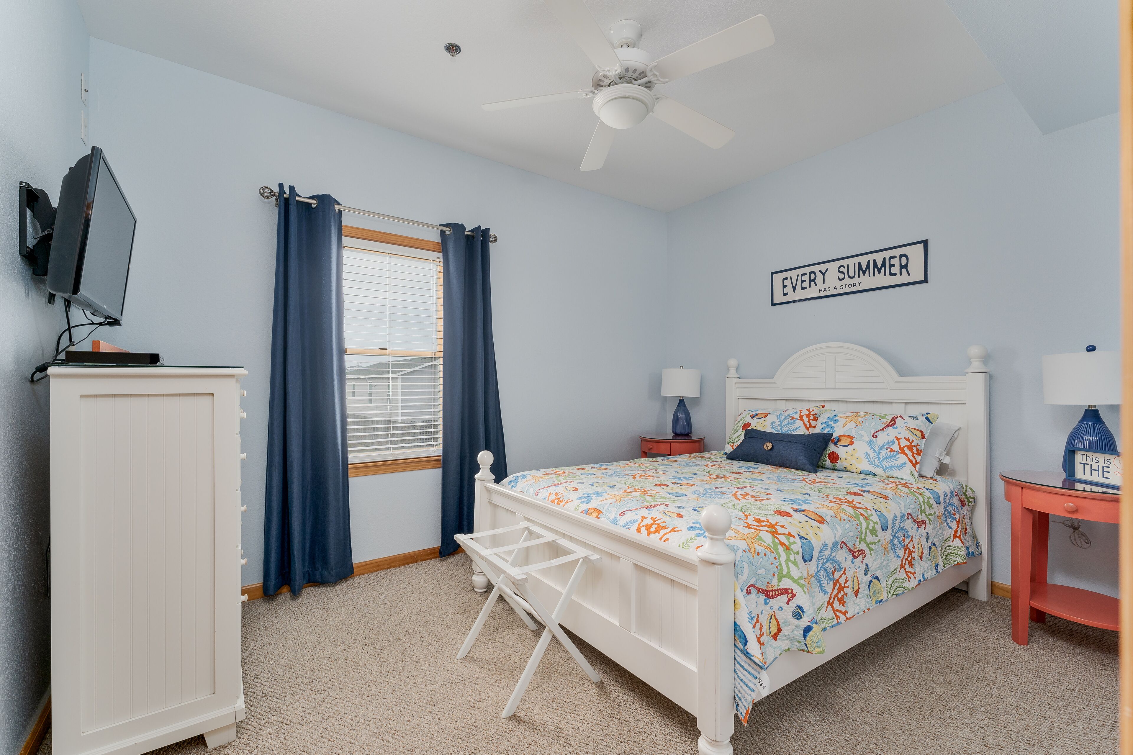 A bedroom with a bed, a ceiling fan, a TV, a window with curtains, and a sign on the wall.