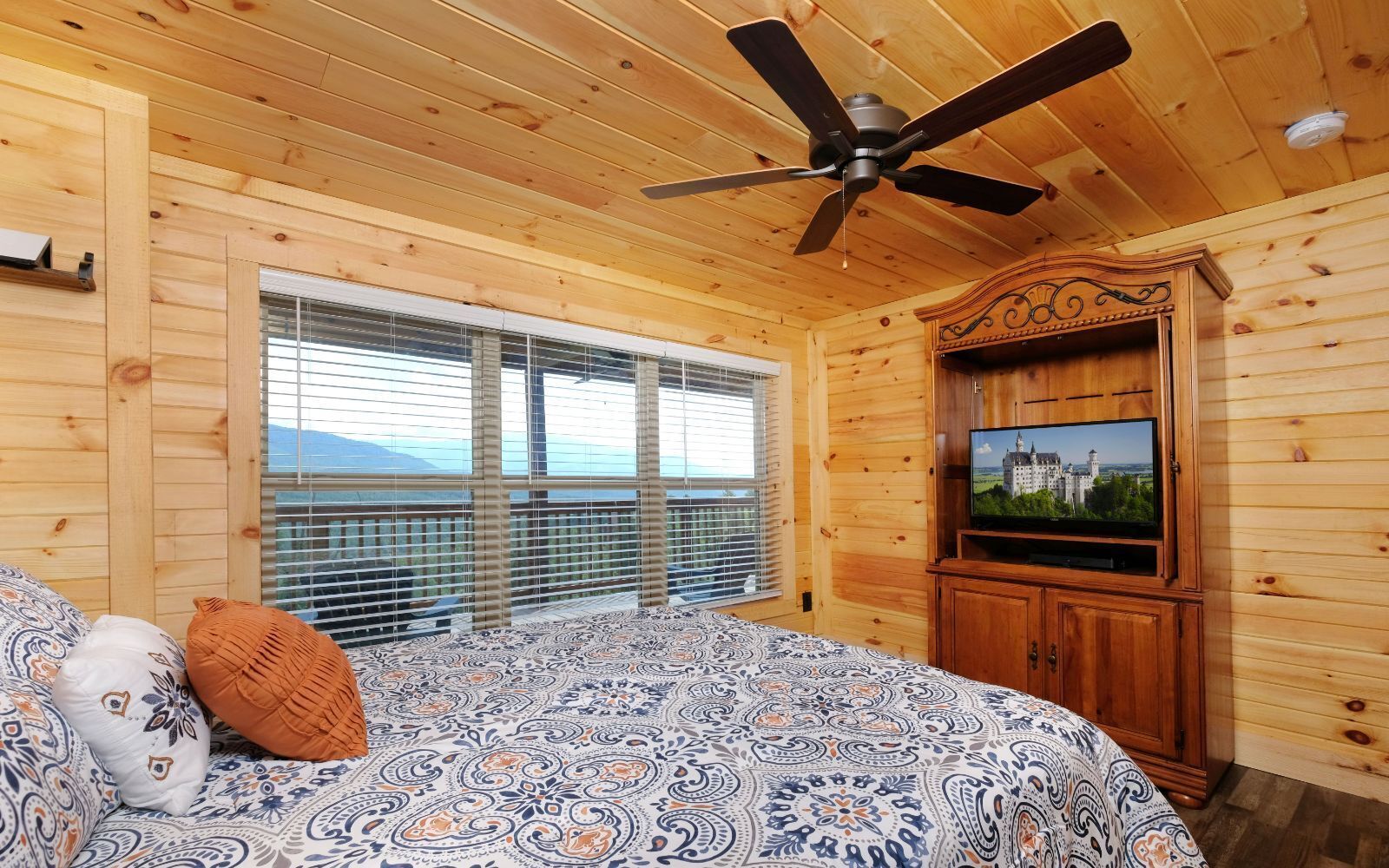 Habitación de cabaña de madera con cama, ventilador de techo, televisor y ventanas amplias con persianas.