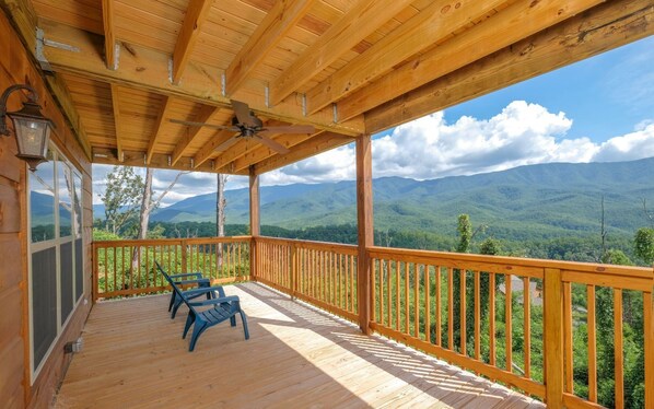 Une terrasse en bois avec un ventilateur de plafond, deux chaises bleues et une vue sur les montagnes.