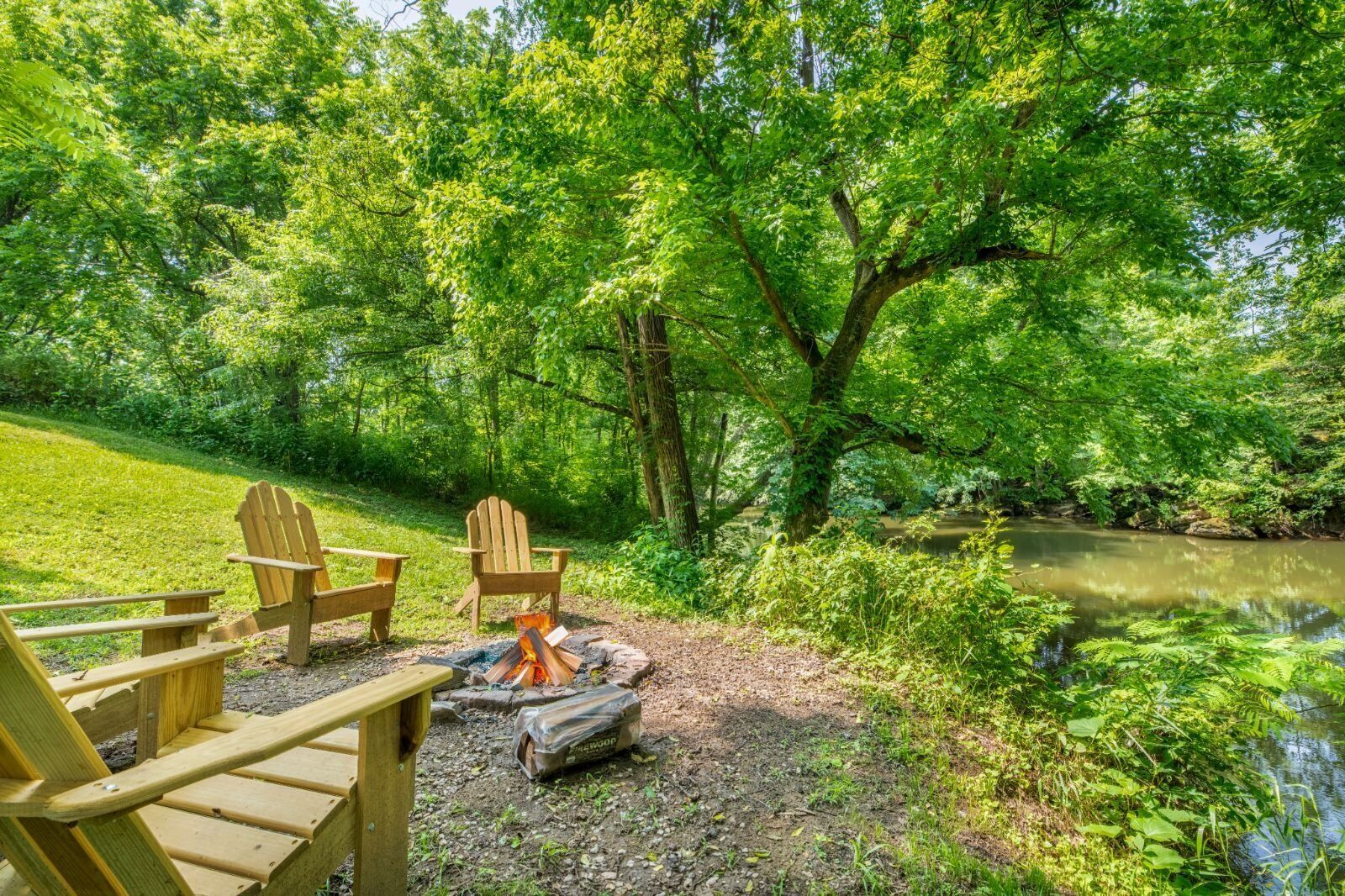 A campsite with wooden chairs, a fire pit, and a river in a forested area.