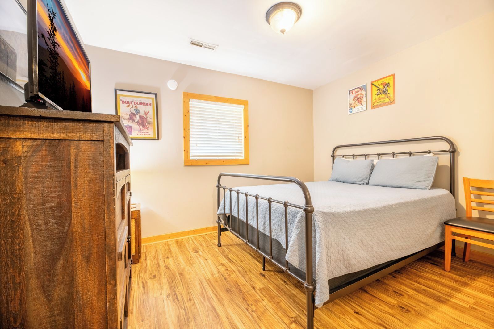 A bedroom with a metal bed frame, a wooden dresser, a window with blinds, and a poster on the wall.