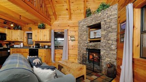 A cozy cabin interior with a stone fireplace, wooden furniture, and a kitchen area.