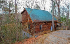 A rustic cabin with a metal roof, surrounded by trees and a gravel driveway.