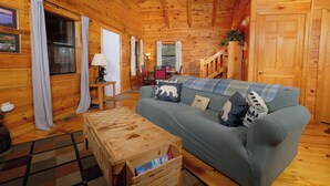 A cozy cabin interior with a wooden floor, a sofa with plaid cushions, a wooden coffee table, and a staircase leading to an upper level.
