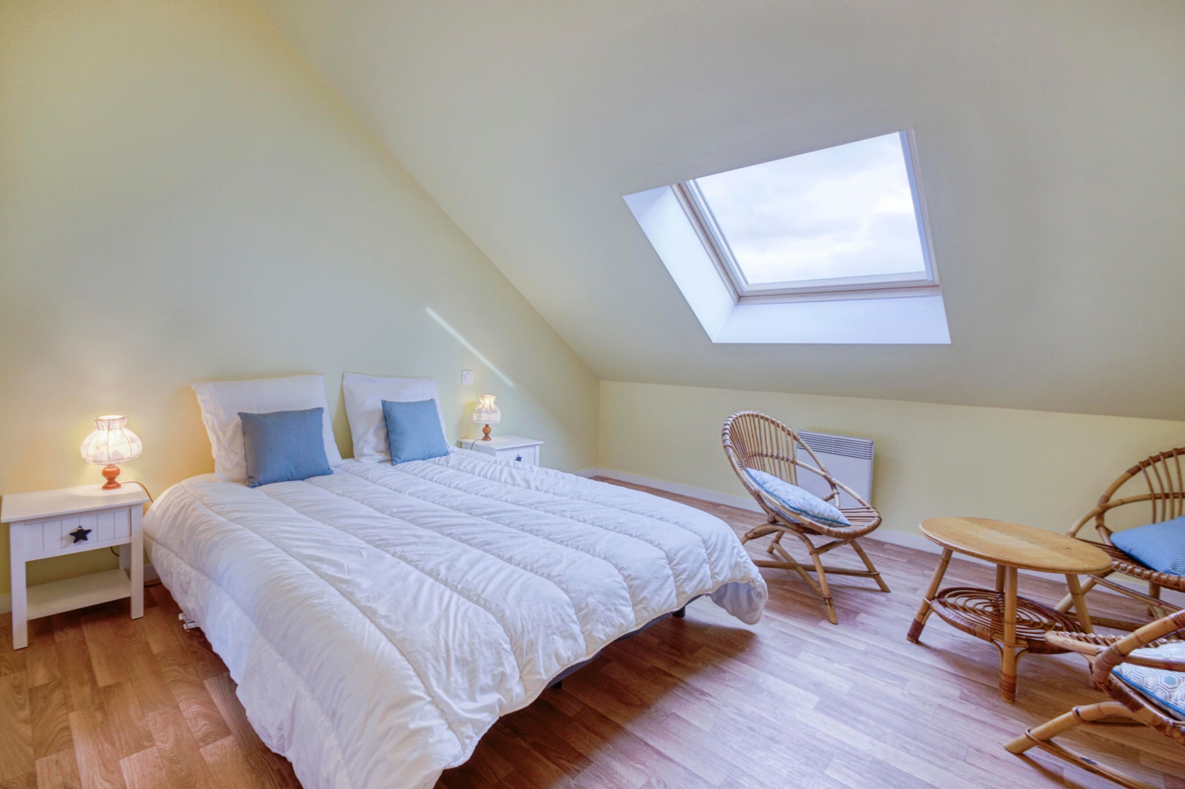 Quarto com teto inclinado, claraboia, cama com roupa de cama branca, duas mesas de cabeceira com luminárias e móveis de vime.