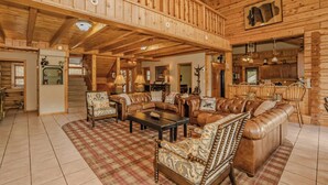 A spacious log cabin interior with a high ceiling, a central seating area with leather sofas, a dining area with wooden chairs, and a kitchen visible through an open doorway.