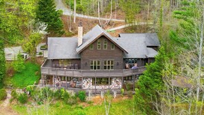 A large log cabin with a stone chimney, a covered deck with outdoor seating, and a scenic forest backdrop.