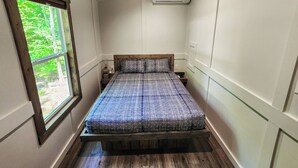 Une petite chambre avec un plafond en bois, une fenêtre donnant sur la verdure, un lit recouvert d’une couvre-lit à motifs et un climatiseur fixé au mur.