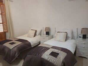 Un dormitorio con dos camas individuales, cada una con una mesita de noche y una lámpara. La habitación tiene una puerta de madera y una ventana con persianas.