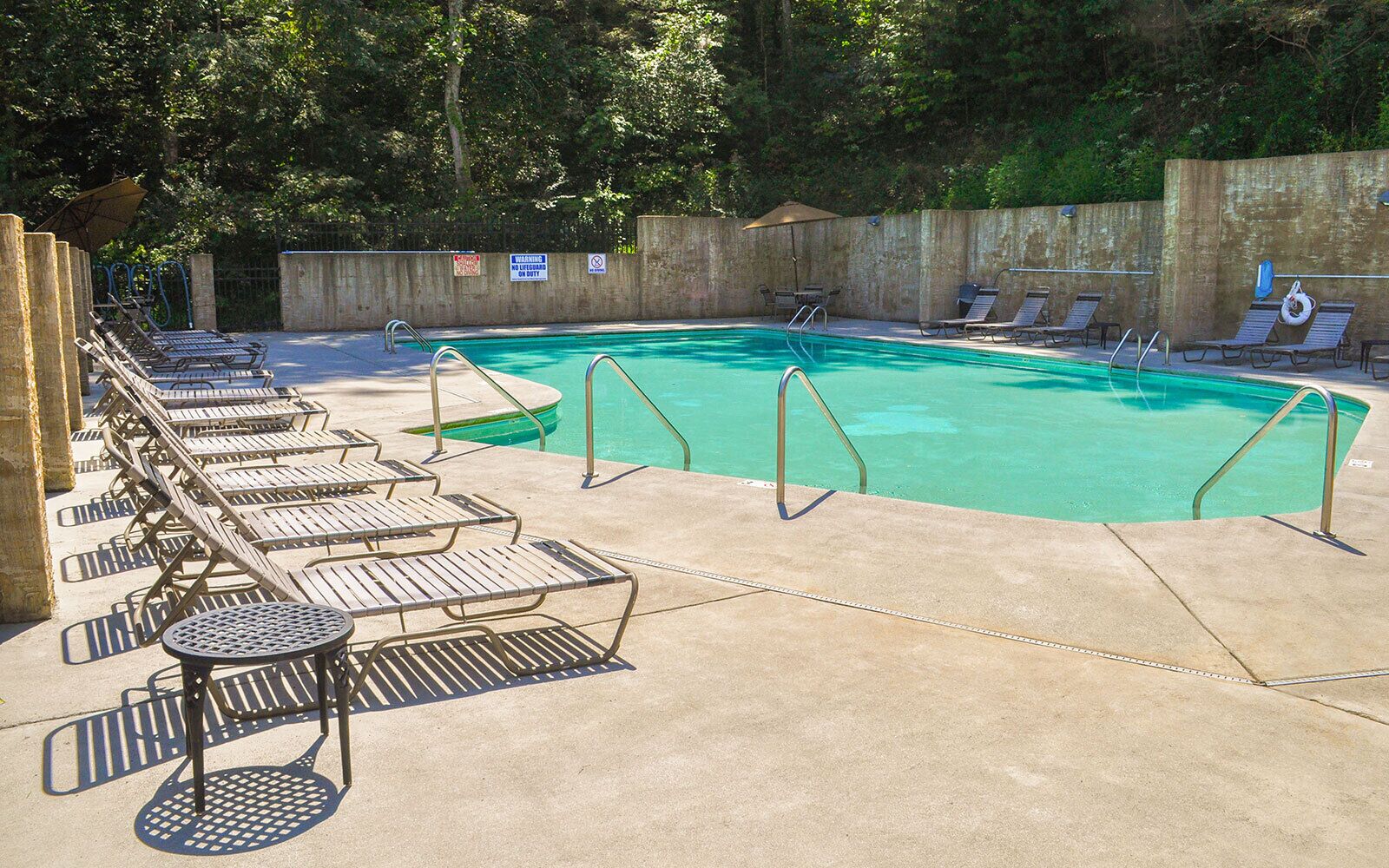 A swimming pool with lounge chairs and a table, surrounded by a concrete deck and a wooden fence.