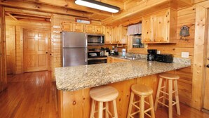 A kitchen with wooden cabinets, a granite countertop, stainless steel appliances, and wooden stools. - Mountain Time – 5BR Game Room & Hot Tub Cabin, Eagle Ridge Resort (Pigeon Forge)