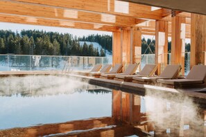 Una piscina infinita de madera con sillas de descanso, rodeada por una terraza de madera y balaustrada de vidrio, con vistas a una zona montañosa cubierta de bosque.