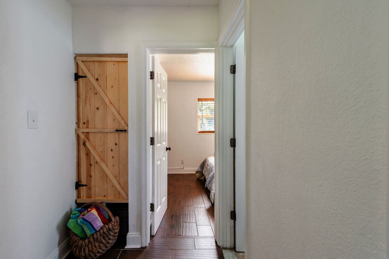 A hallway with a wooden barn-style door, a basket with colorful items, and a bed with a window in the background.
