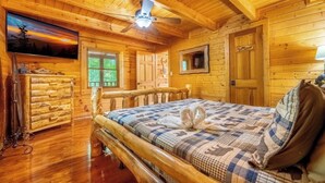 Una habitación de cabaña de troncos con una cama, paredes de madera, un ventilador de techo, un aparador y una ventana con persianas.