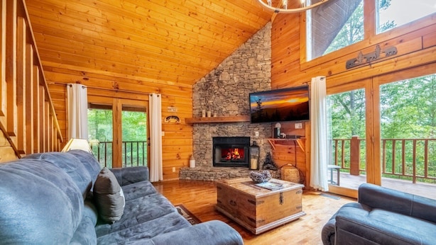 Interior acogedor de una cabaña con chimenea de piedra, paredes de madera y un amplio ventanal con vistas a un bosque.
