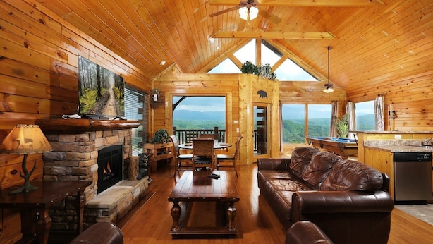 L’intérieur chaleureux d’un chalet avec une cheminée en pierre, un canapé en cuir, une table à manger en bois et une vue sur les montagnes par de grandes fenêtres.
