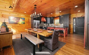 A modern kitchen with wooden cabinets, a stainless steel refrigerator, and a dining area with a wooden table and bench. - Point of Grace - Private Indoor Pool, hot tub, theater room, game room, mountain views, & more! (Pigeon Forge)