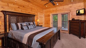 A wooden cabin bedroom with a large bed, a ceiling fan, a dresser, and a TV mounted on the wall.