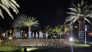 Un espace extérieur aménagé pour la nuit, avec des palmiers, un mur en pierre et un bâtiment dont les fenêtres sont illuminées.