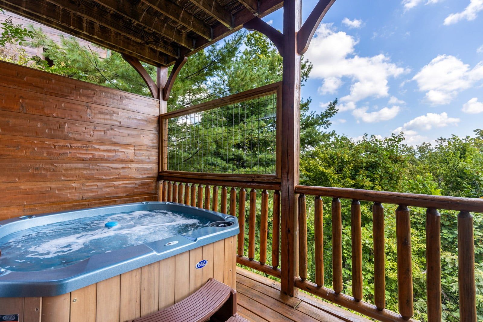 Una terraza de madera con un jacuzzi, barandas de madera y vistas a un bosque.