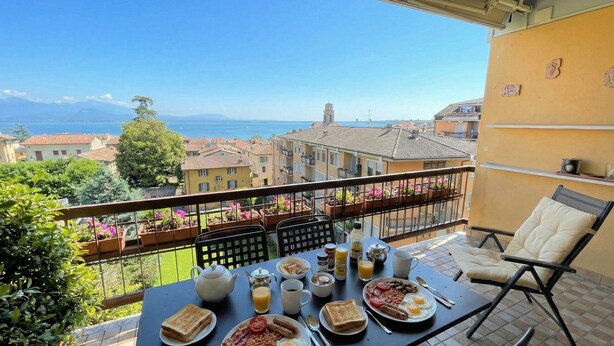 A balcony with a table set for breakfast, overlooking a scenic view of buildings and the sea. - DesenzanoLoft Ciao Lago  (Desenzano del Garda)
