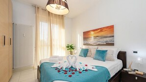 A hotel room with a bed, a painting of a sunset, a potted plant, and a nightstand with a lamp. - DesenzanoLoft Ciao Lago  (Desenzano del Garda)