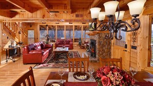 A spacious log cabin interior with a fireplace, leather sofas, wooden dining table, and a chandelier.