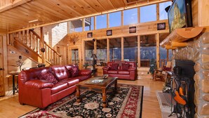 A spacious log cabin interior with a large living area featuring leather sofas, a coffee table, and a fireplace.