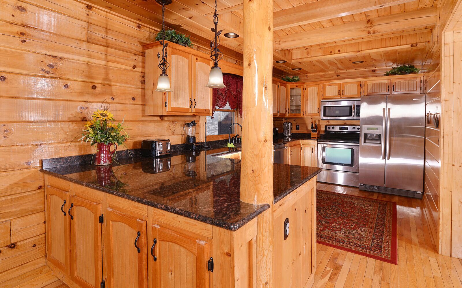 Interior de una cabaña de madera con cocina, que incluye una encimera de granito, muebles de madera y electrodomésticos de acero inoxidable como un refrigerador y un microondas.