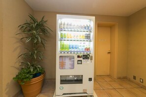 Máquina expendedora con diversas bebidas y aperitivos, una planta en maceta y una puerta al fondo.