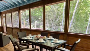 Comedor con mesa de madera preparada para cuatro personas, rodeada de sillas de mimbre con cojines. Amplias ventanas ofrecen vista a un área boscosa.