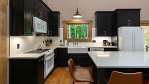Una cocina moderna con armarios oscuros, encimera blanca, horno integrado, microondas y ventana con vistas a árboles.