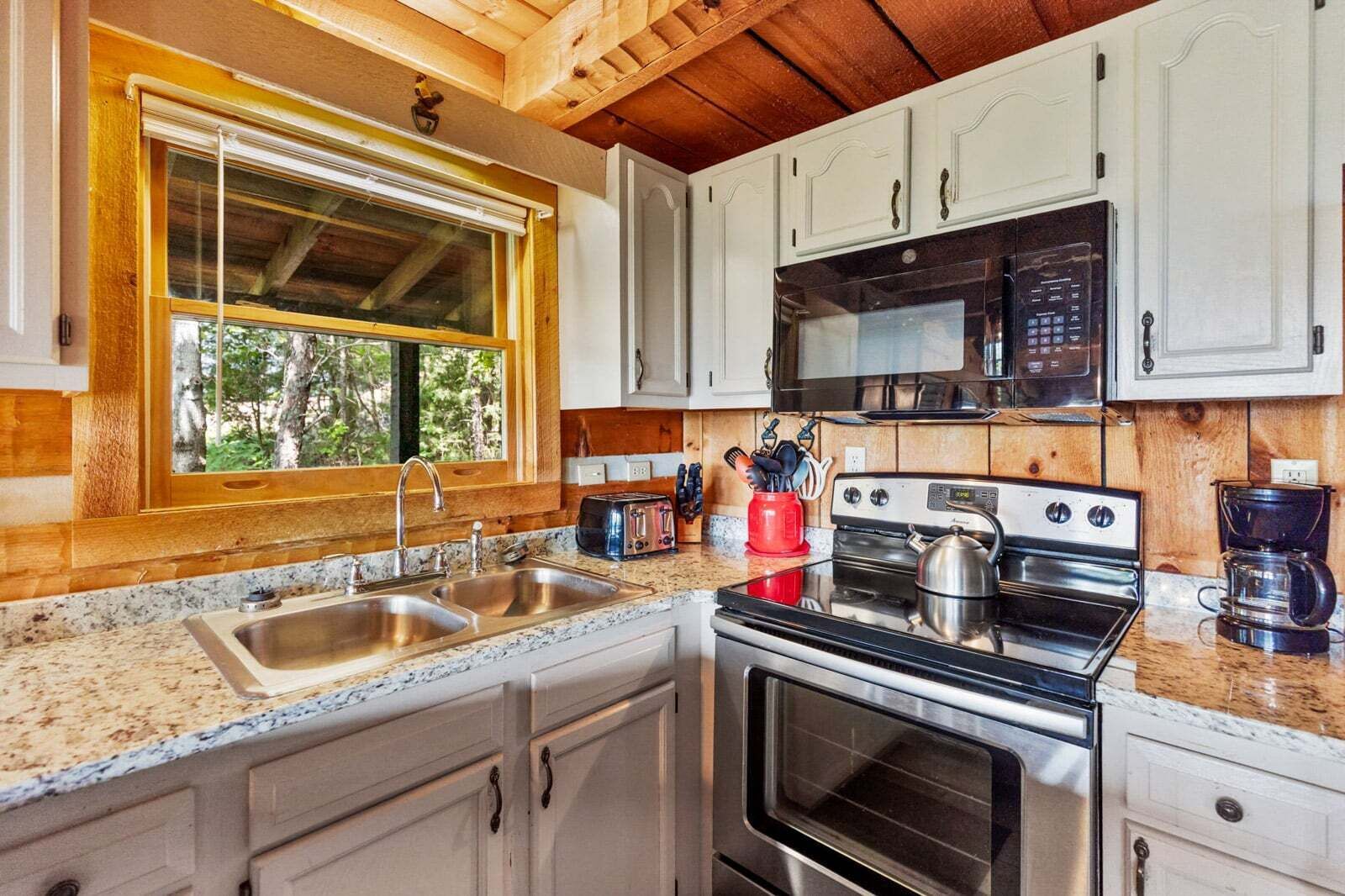 A kitchen with a double sink, a microwave, a stove, and a window with a view of trees.